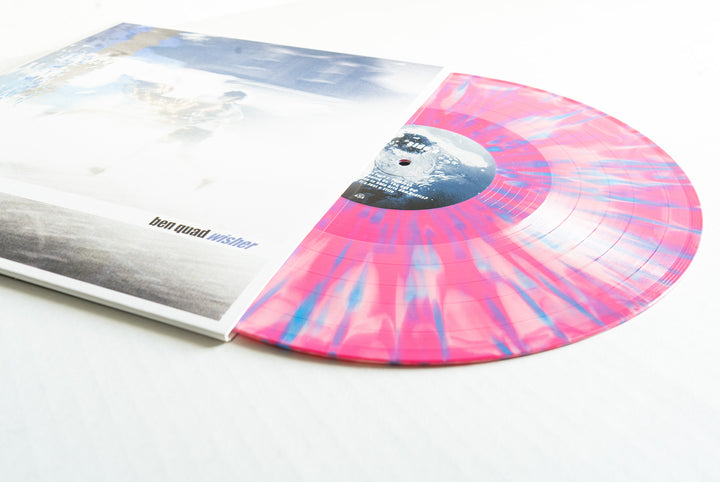A Baby Pink Blue Splatter Wisher Vinyl by Ben Quad partially slides from a white album cover with blurred artwork and "ben quad wistler" on the front, resting on a clean white surface—ideal for an album presale display.