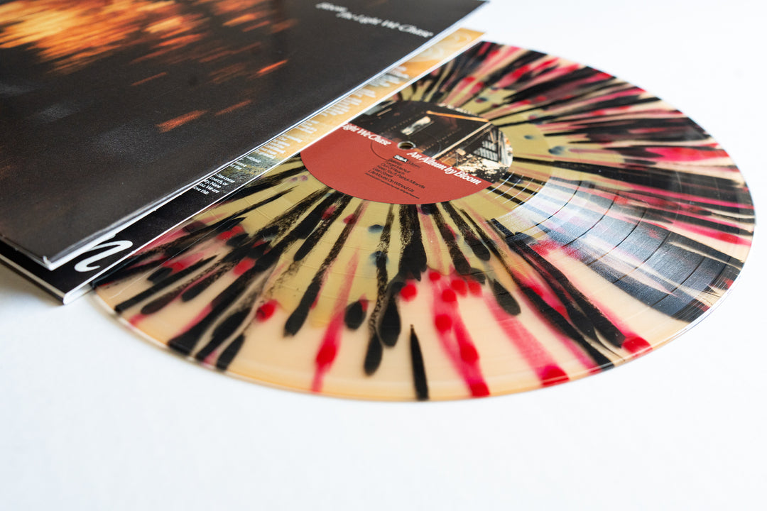 A vibrant Gold In Beer vinyl from Bloom, with black and red splatters on a clear base, slides out of its album cover showcasing a warm-toned, blurry image. The brown center label features crisp white Bloom branding.