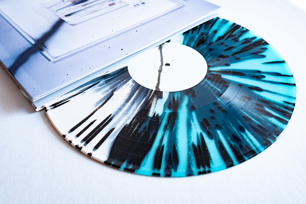 A blue and heavy black splatter-patterned Desertion Vinyl LP by Spite House partially slides out from a white minimalist album cover, resting on a white surface. Album release 2025.