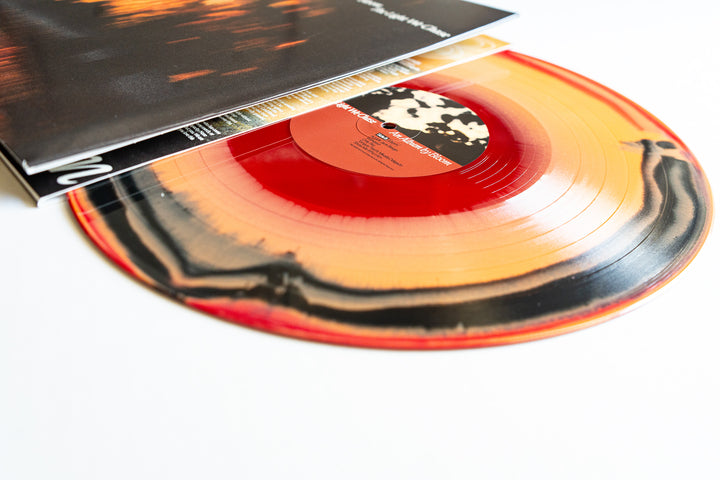 A limited edition Bloom multicolored vinyl record with red, orange, black, and yellow swirls partially slides out from its album cover on a white surface.