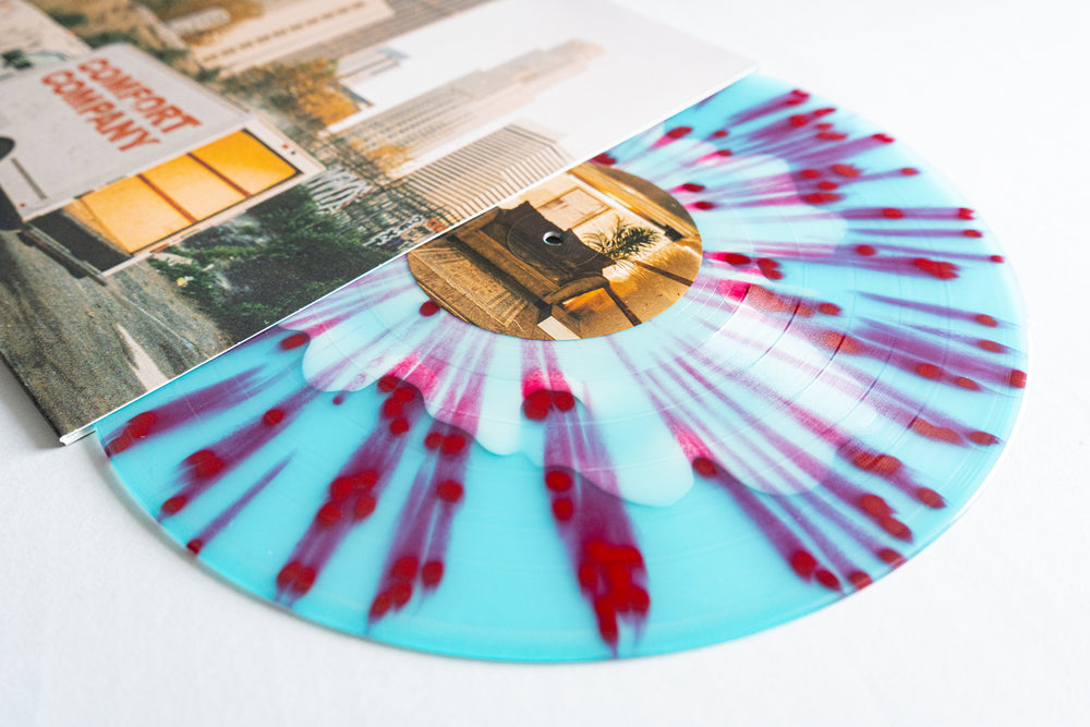 An Electric Blue Vinyl LP by Bearings, featuring red and white splatter, partially slides out from an album cover that shows an urban city scene and a “Comfort Company” truck.