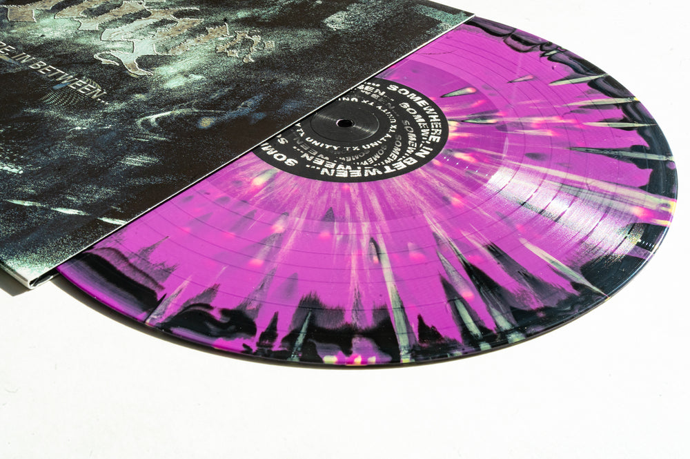 A UNITYTX Somewhere In Between LP Black and Purple Splatter Vinyl partially slides from its sleeve, showing distressed white text on a dark background. The vinyl presale label is visible at the center.