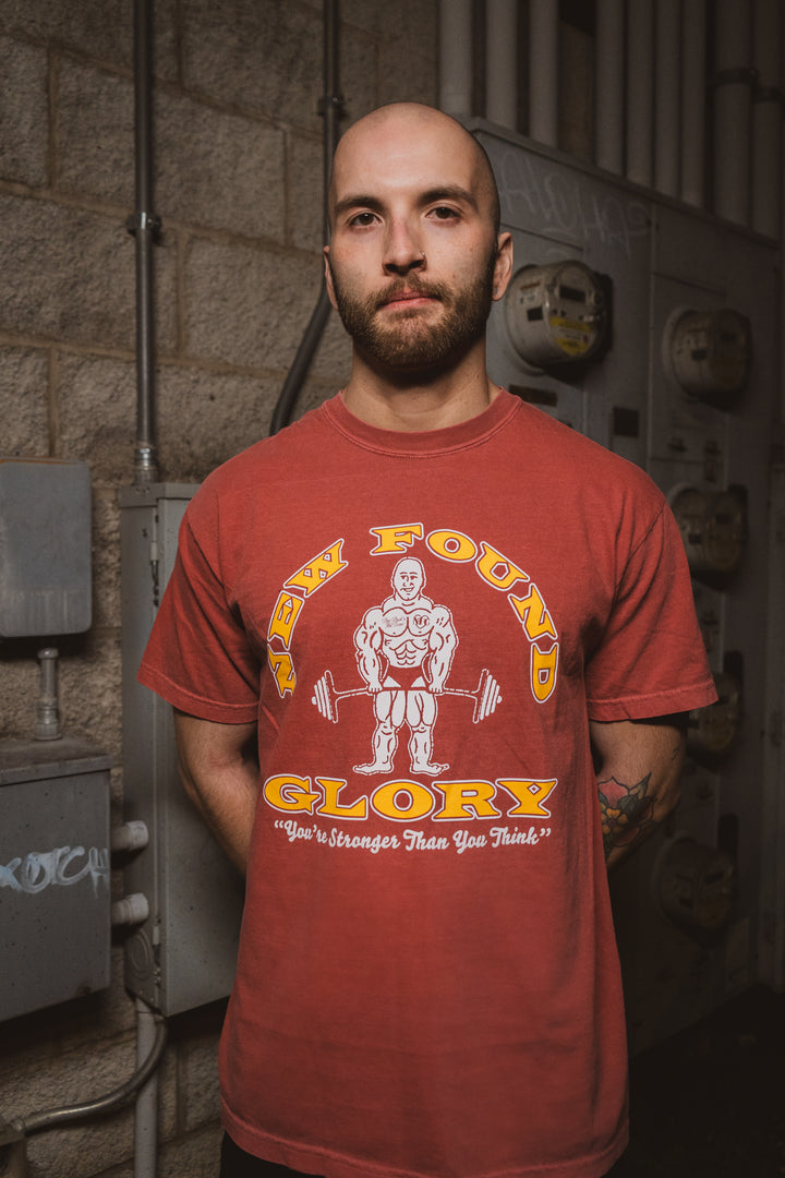 A man with a shaved head and beard stands by utility meters, wearing a red New Found Glory t-shirt with a cartoon bodybuilder and the text “You’re stronger than you think.”.