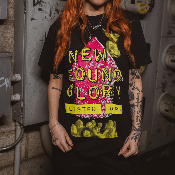 A person with long red hair and tattoos wears a New Found Glory Comfort Colors black shirt featuring a bold pink and yellow “New Found Glory” and “Listen Up!” graphic; electrical equipment appears in the background.