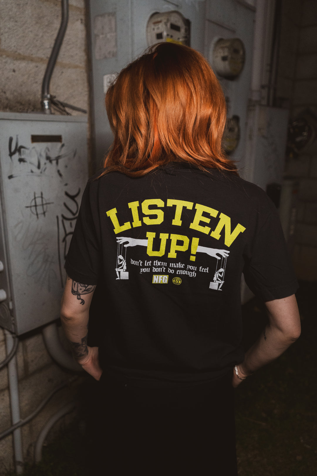 A person with long red hair faces away, wearing a black New Found Glory Comfort Colors shirt featuring bold yellow "LISTEN UP!" text above smaller white graphics. Utility boxes and concrete walls appear in the background.