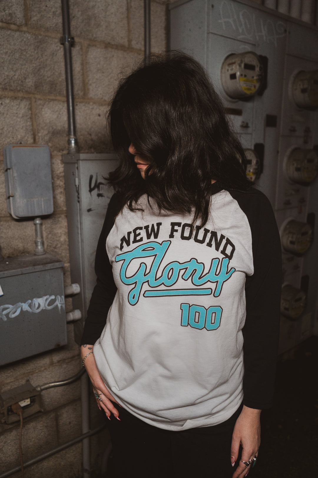 A person with shoulder-length dark hair looks down, wearing a New Found Glory black and white 3/4 sleeve Raglan Baseball T-shirt, standing in front of a wall with electrical meters and graffiti.