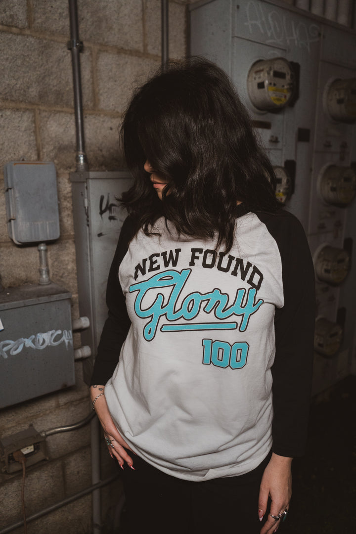 A person with shoulder-length dark hair looks down, wearing a New Found Glory black and white 3/4 sleeve Raglan Baseball T-shirt, standing in front of a wall with electrical meters and graffiti.