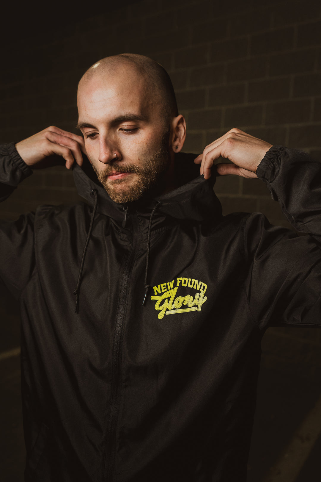 A man with a shaved head adjusts the hood of his black New Found Glory jacket, featuring the yellow band logo on the chest, while standing in front of a dark brick wall.
