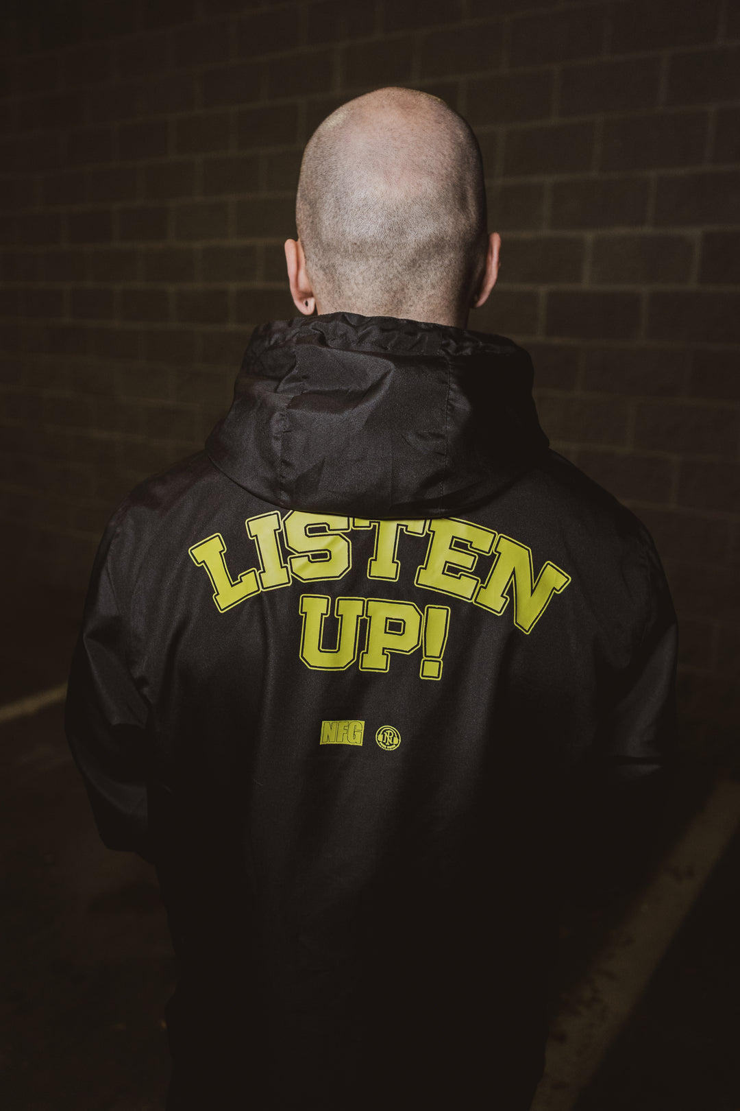 A person with a shaved head, seen from behind, wears a black New Found Glory jacket with “LISTEN UP!” in bold yellow on the back, standing before a brick wall in dim lighting.