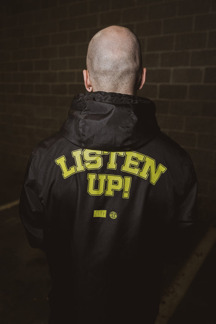 A person with a shaved head, seen from behind, wears a black New Found Glory jacket with “LISTEN UP!” in bold yellow on the back, standing before a brick wall in dim lighting.
