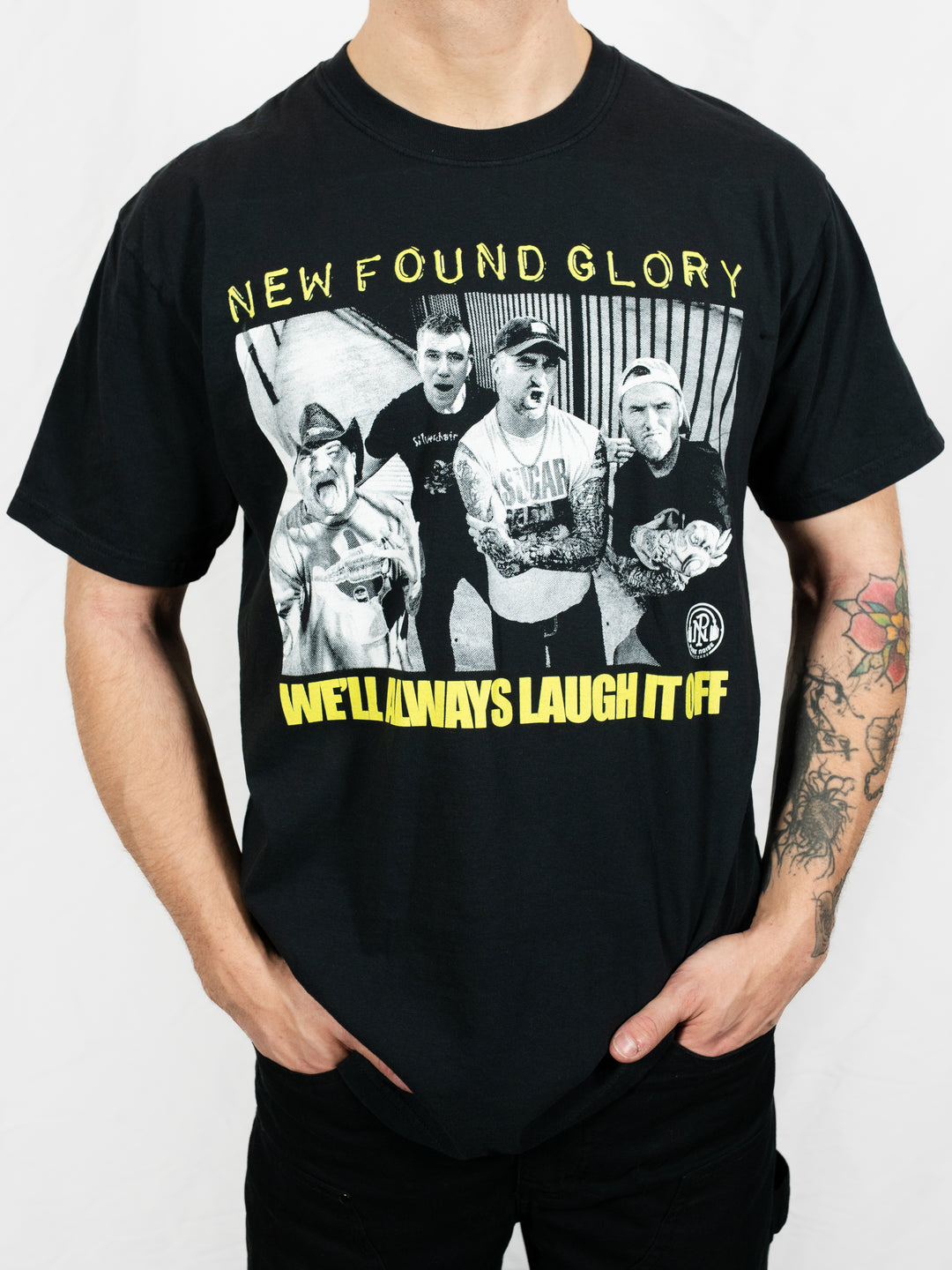 A person wears a black New Found Glory Comfort Colors t-shirt with a black-and-white photo of four band members and yellow text reading "NEW FOUND GLORY" and "WE'LL ALWAYS LAUGH IT OFF," showing tattooed arms.