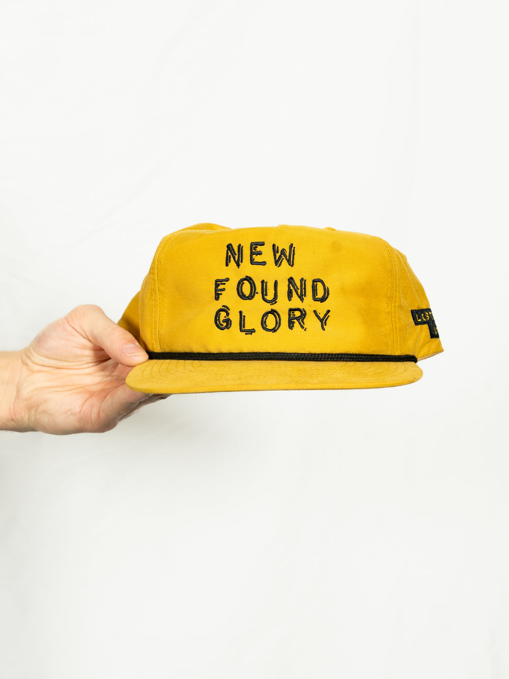 A hand holds a yellow New Found Glory snapback cap with the band's name embroidered in black on the front, set against a plain white background.