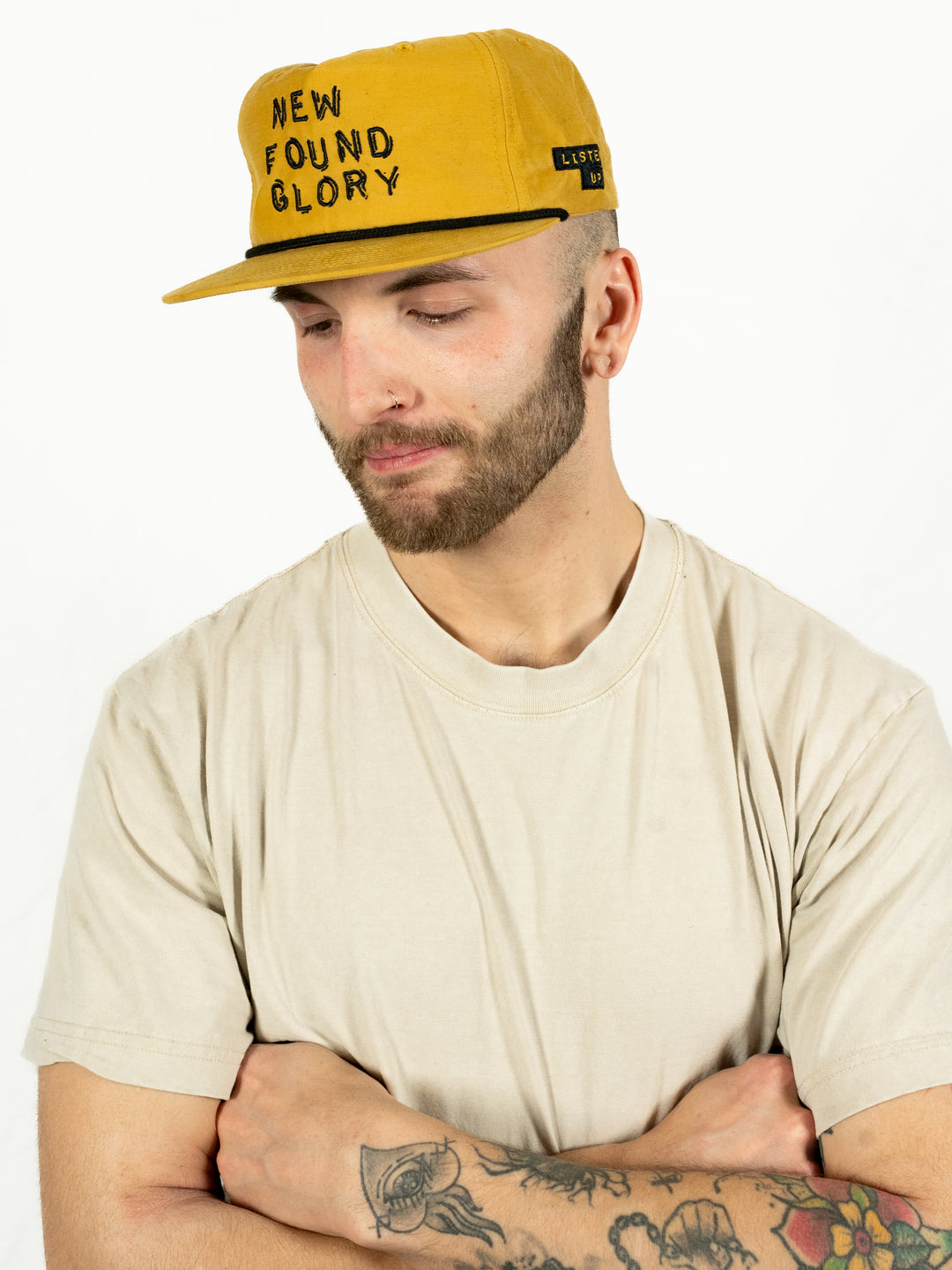 A man in a beige t-shirt and a mustard yellow New Found Glory Richardson Umpqua Snapback hat stands against a white background, looking down with arms crossed. He has a beard, earrings, and tattoos on his forearm.