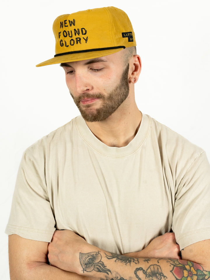 A man in a beige t-shirt and a mustard yellow New Found Glory Richardson Umpqua Snapback hat stands against a white background, looking down with arms crossed. He has a beard, earrings, and tattoos on his forearm.
