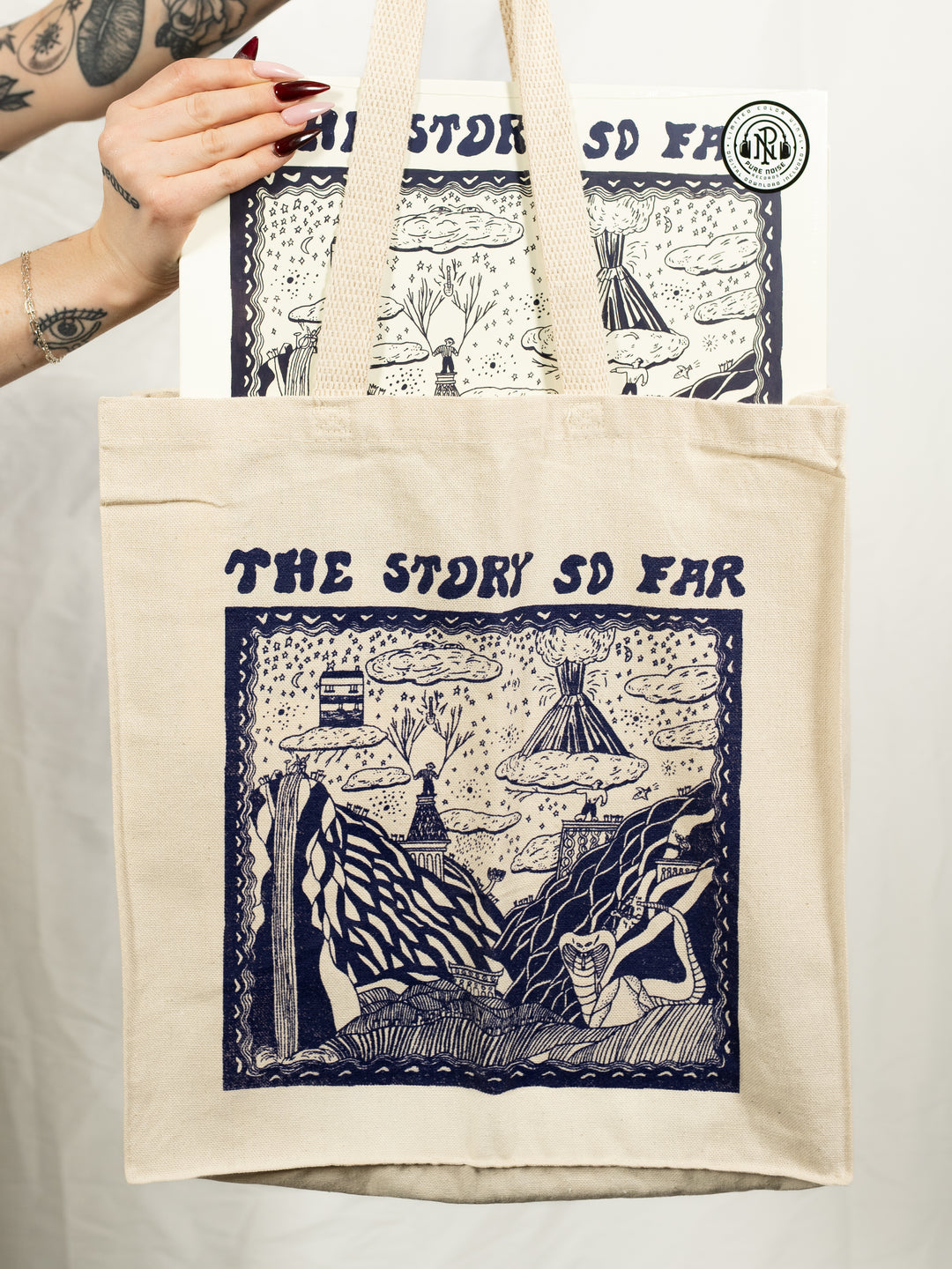 A tattooed individual holds a natural tote bag and matching print, both featuring blue album art and "The Story So Far" text above surreal landscape artwork.