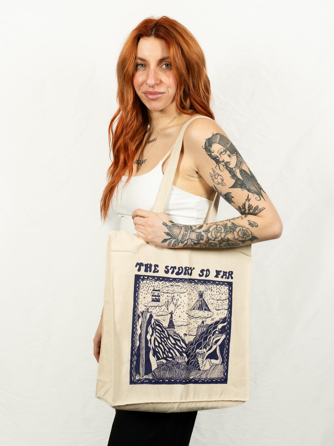 A woman with long red hair and tattoos smiles while holding a natural tote bag from The Story So Far, featuring blue illustrated album art and the band’s name. She stands against a plain white background.