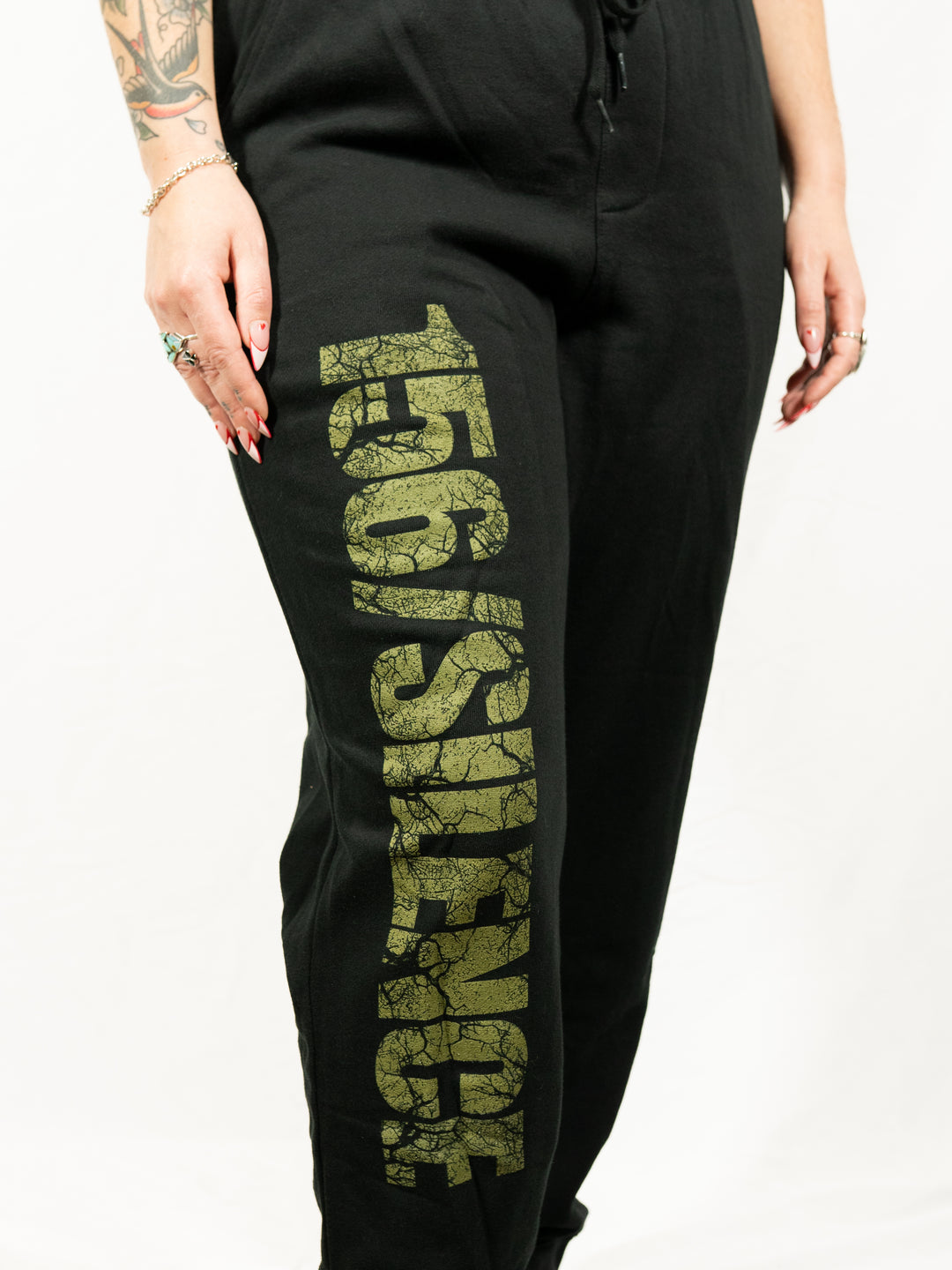 A person with tattoos, rings, and a bracelet wears black 156/Silence midweight fleece sweatpants by Independent Trading Co., featuring large textured gold "156/SILENCE" lettering down the leg. The background is white.