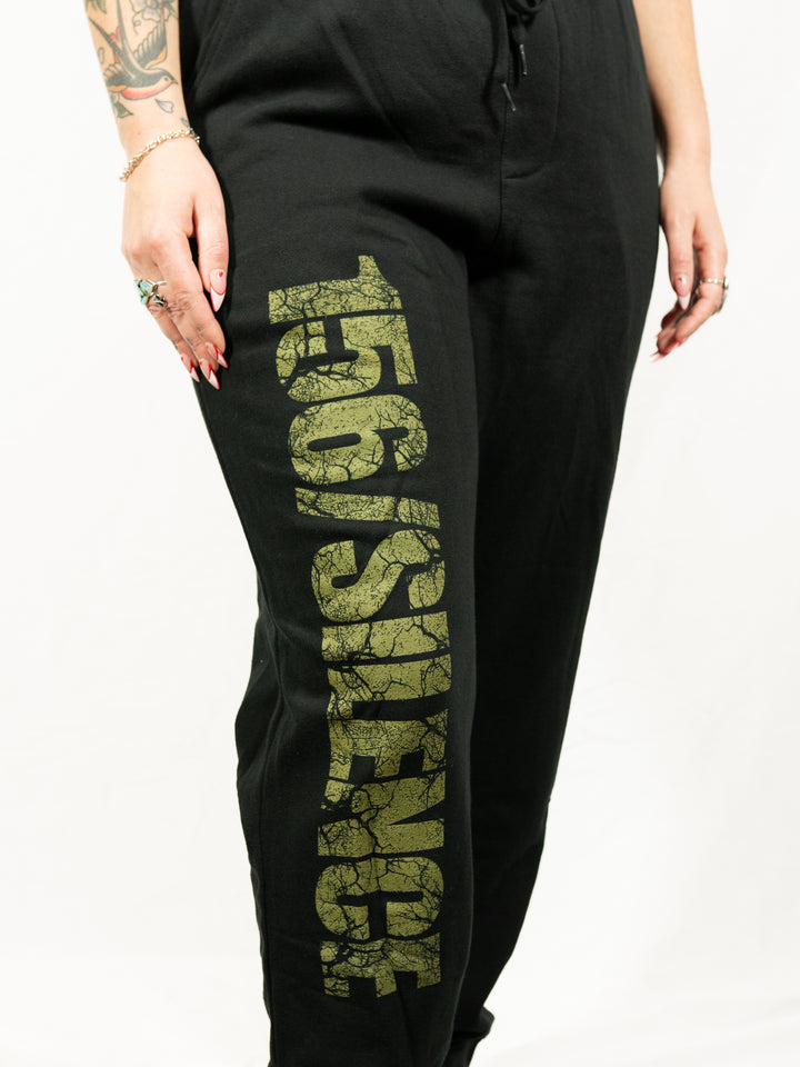 A person with tattoos, rings, and a bracelet wears black 156/Silence midweight fleece sweatpants by Independent Trading Co., featuring large textured gold "156/SILENCE" lettering down the leg. The background is white.