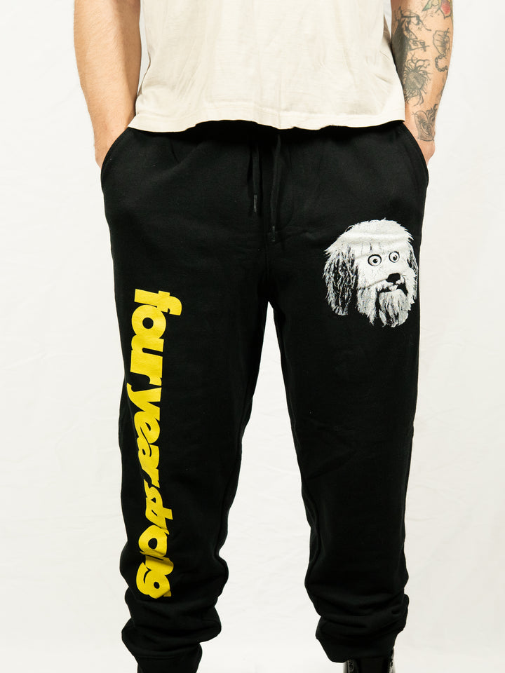 A person wearing black Four Year Strong fleece sweatpants featuring "FOUR YEAR STRONG" in bold yellow letters down one leg and a cartoon dog face on the other, standing with hands in pockets.