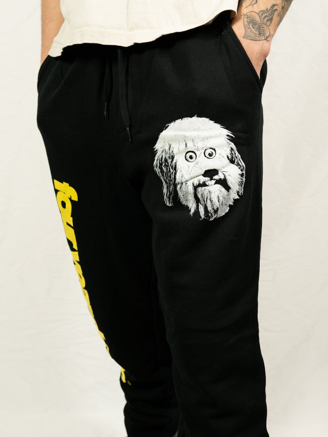 Person wears black midweight fleece sweatpants from Four Year Strong, featuring a white and gray cartoon dog face on the upper thigh and yellow text along the leg, standing against a plain white background. Only lower torso and legs are visible.