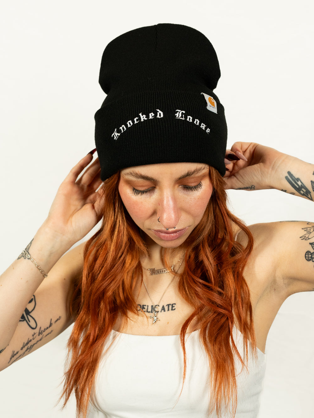 A woman with long red hair and tattoos adjusts her black embroidered Knocked Loose cuffed beanie, featuring white text. She pairs it with a white sleeveless top against a plain background.
