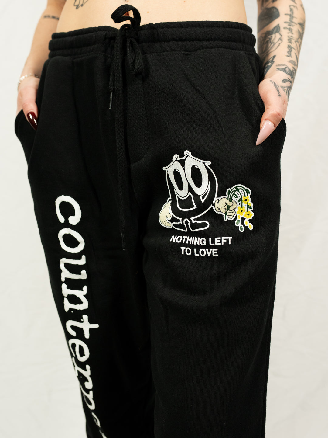 A person wearing black Counterparts sweatpants—"Counterparts" printed vertically on one leg, a cartoon Sad Face with "NOTHING LEFT TO LOVE" on the other—poses with tattooed hands in pockets, paired with a black T-shirt.