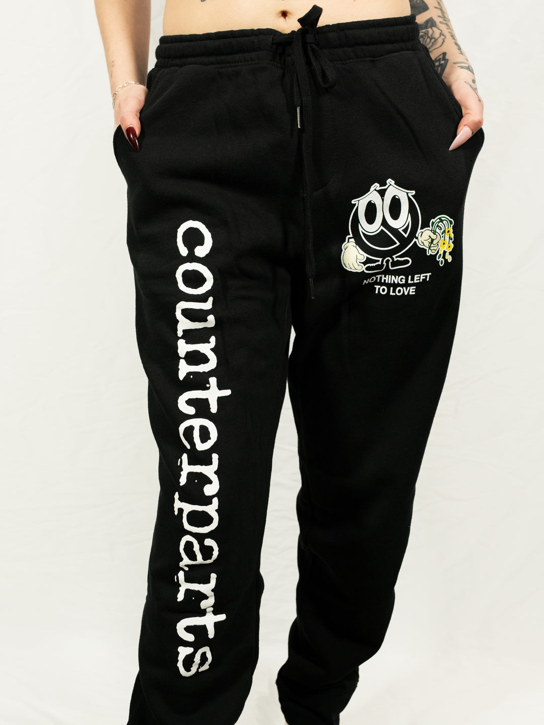 A person wears Counterparts black sweatpants featuring "COUNTERPARTS" in bold white lettering down one leg and a Sad Face graphic with "NOTHING LEFT TO LOVE" near the pocket, hands tucked casually in the pockets.