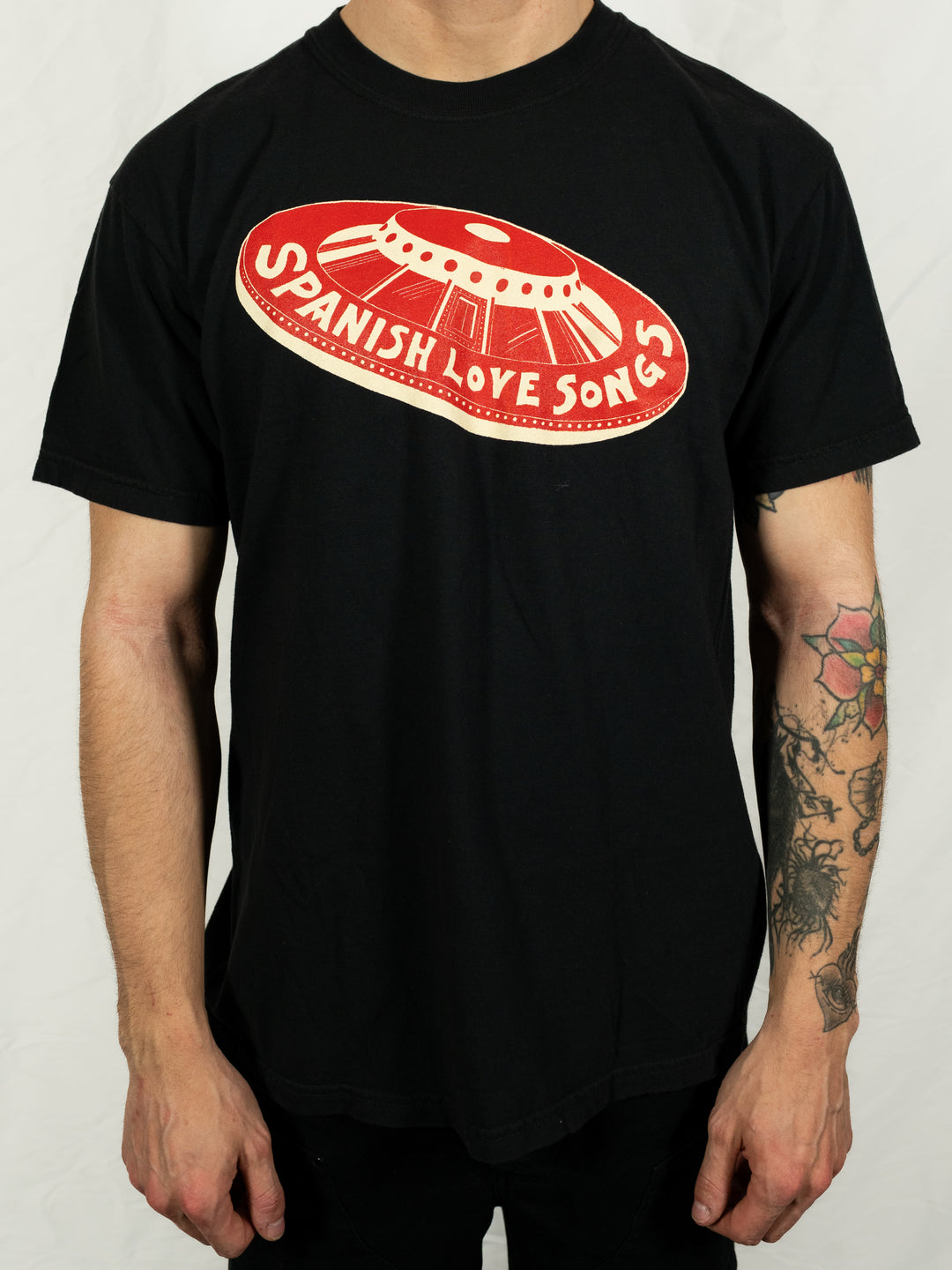 A person wears a black Spanish Love Songs tee featuring a red and white UFO graphic and the band name on the chest. Their tattooed arms are visible.