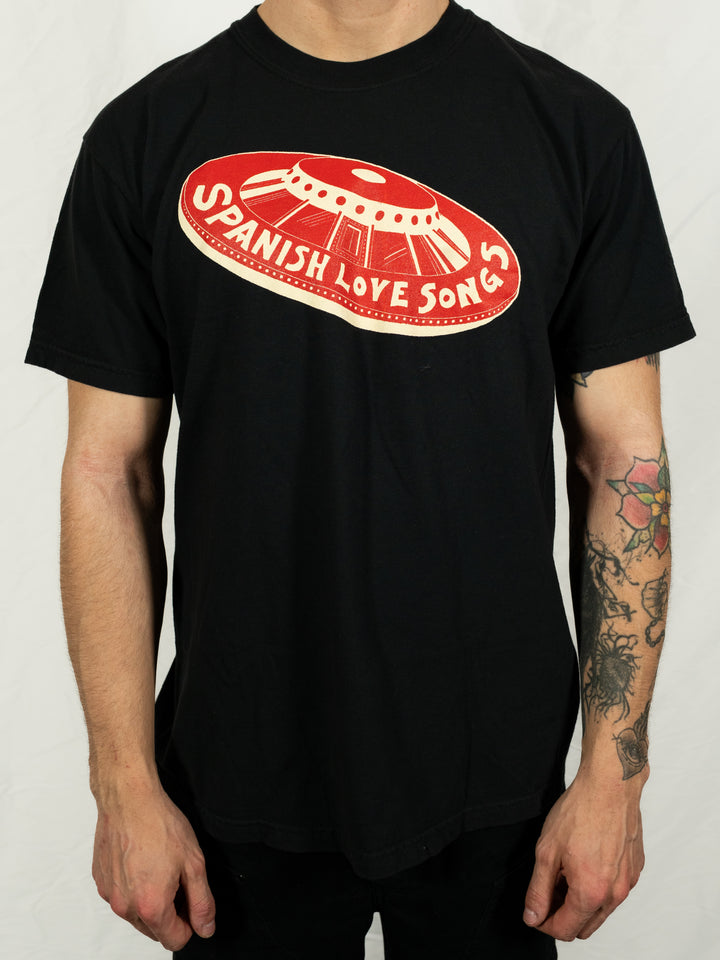 A person wears a black Spanish Love Songs tee featuring a red and white UFO graphic and the band name on the chest. Their tattooed arms are visible.