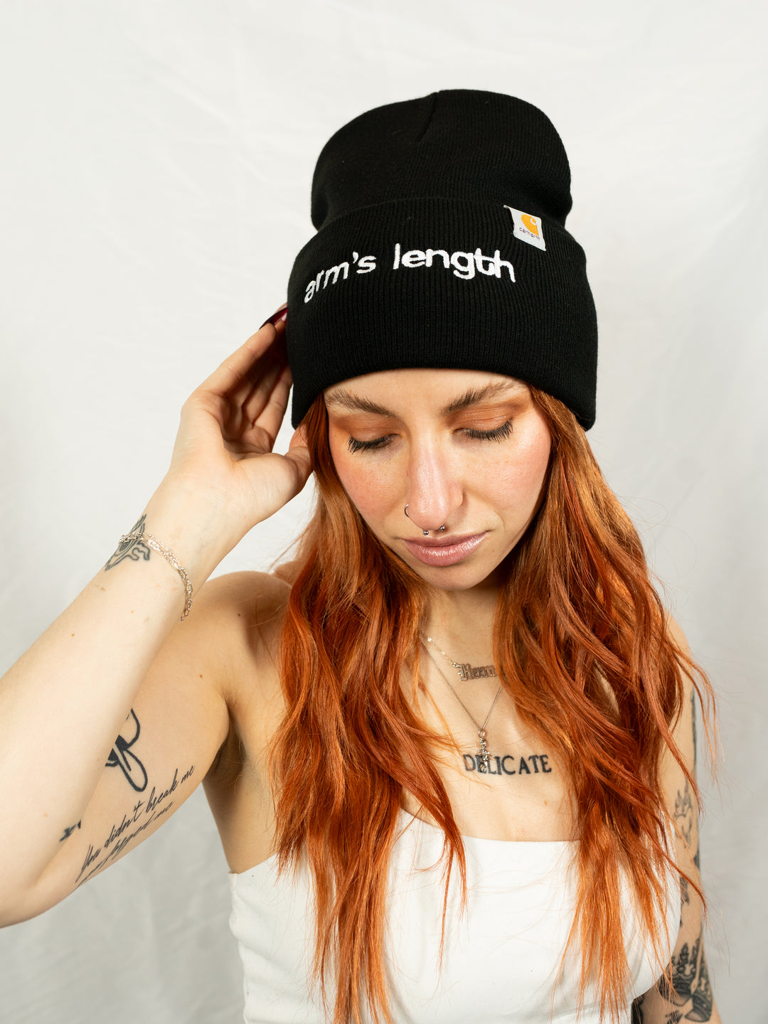 A woman with long red hair and tattoos, wearing a black Arm's Length cuffed beanie and a white top, looks down slightly while touching the beanie. The background is plain and light.