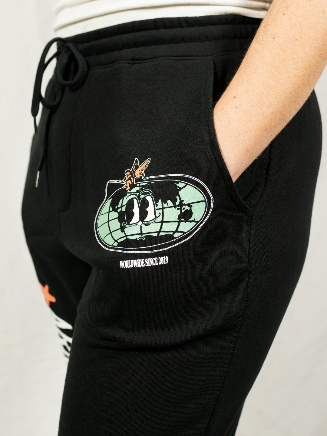 A person wearing black Arm's Length midweight fleece sweatpants with a green and black globe graphic, cartoon eyes, a small orange airplane, and "WORLDWIDE SINCE 2019" on the left pocket area.