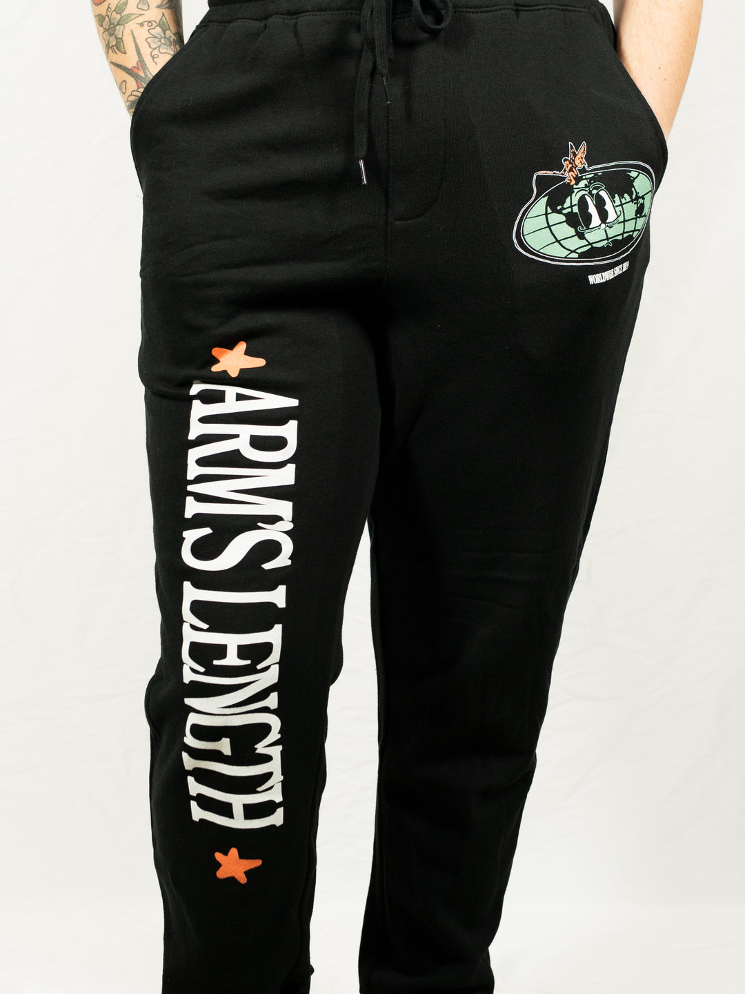 Person wearing black Arm's Length fleece sweatpants with white "ARMS LENGTH" text down one leg, orange star designs, and a planet and rocket patch on the pocket. Only the lower torso and legs are visible.