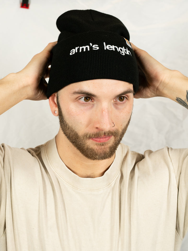 A person wearing a beige t-shirt and a black cuffed beanie featuring the Arm's Length brand adjusts the hat with both hands against a plain white background.