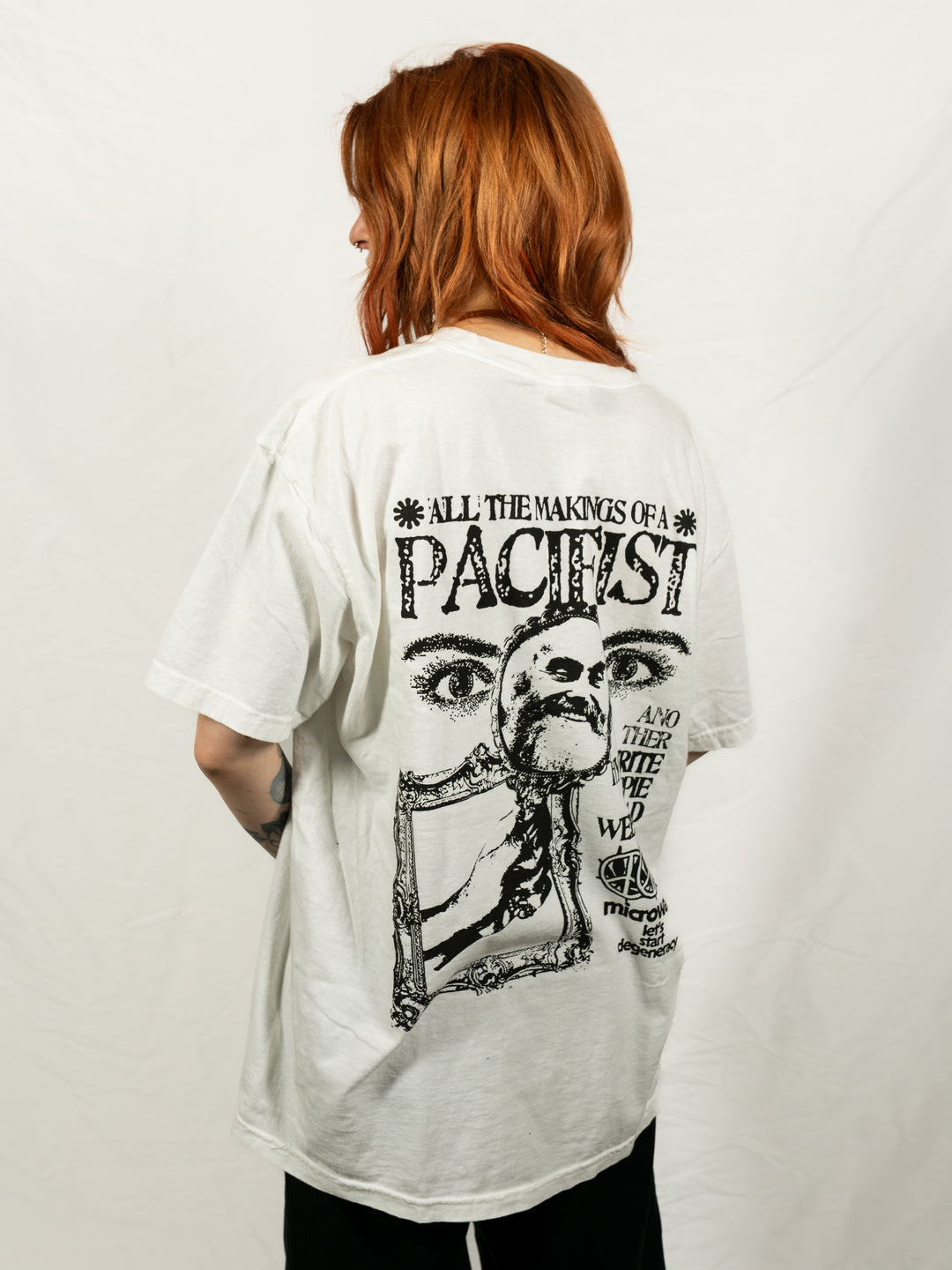 A person with reddish-brown hair, seen from behind, wears a white Microwave T-shirt featuring bold black artwork and the text "ALL THE MAKINGS OF A PACIFIST" above surreal illustrations and additional text.