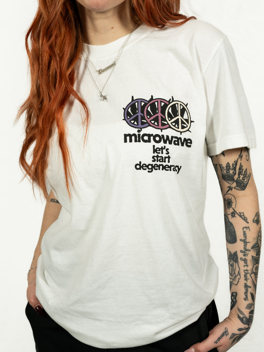 A person with long red hair and tattooed arms wears a white Microwave T-shirt featuring colorful peace signs and the text "lets start degeneracy" on the chest.