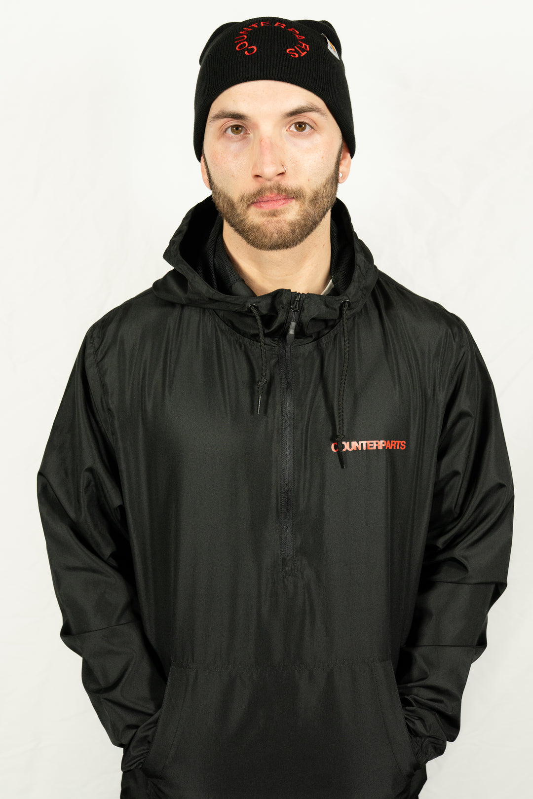 A man stands before a plain white background, wearing a black Counterparts unisex jacket with the brand's name on it and a black beanie with red lettering.