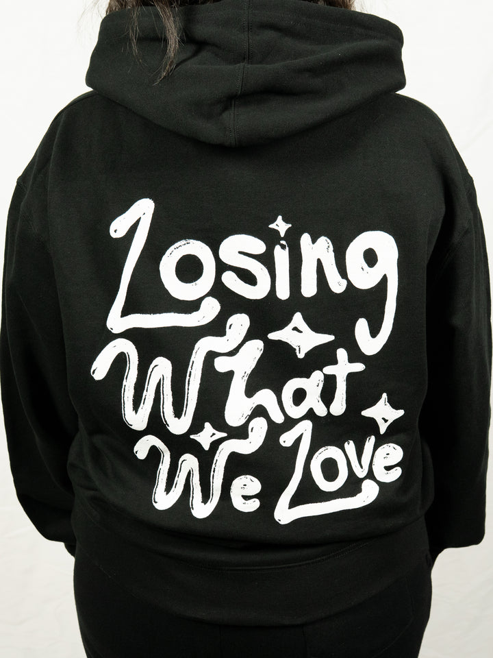 A person wears a black Knuckle Puck hoodie with white handwritten-style "Losing What We Love" text and small star-like accents printed on the back.