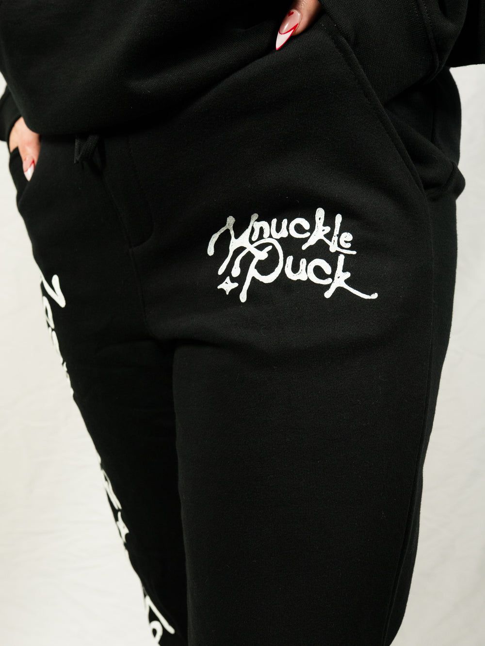A person wears black Knuckle Puck midweight fleece sweatpants with "Knuckle Puck" in white script on the pocket, standing against a plain white background.