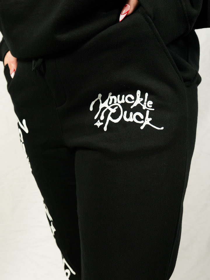 A person wears black Knuckle Puck midweight fleece sweatpants with "Knuckle Puck" in white script on the pocket, standing against a plain white background.