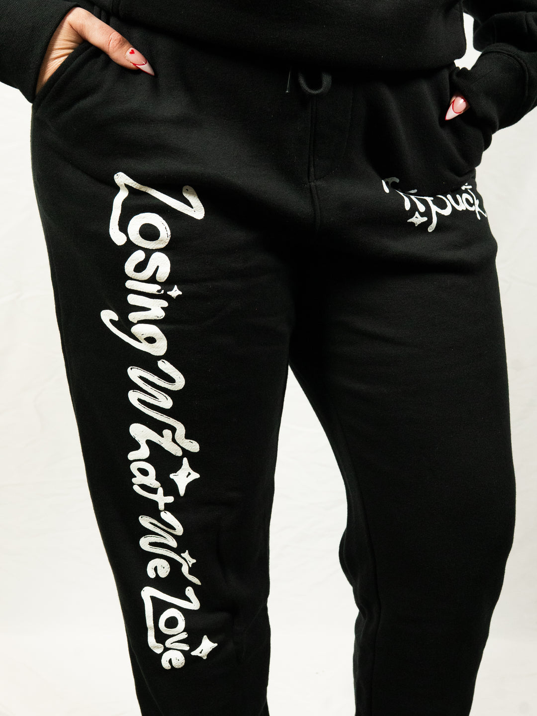 A person stands against a plain white background wearing black midweight fleece sweatpants from Knuckle Puck, featuring white “Losing What We Love” lettering on one leg and partially visible text on the other.