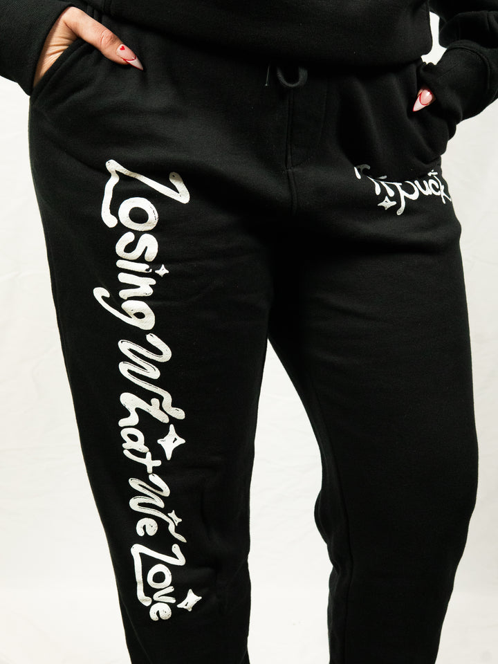 A person stands against a plain white background wearing black midweight fleece sweatpants from Knuckle Puck, featuring white “Losing What We Love” lettering on one leg and partially visible text on the other.
