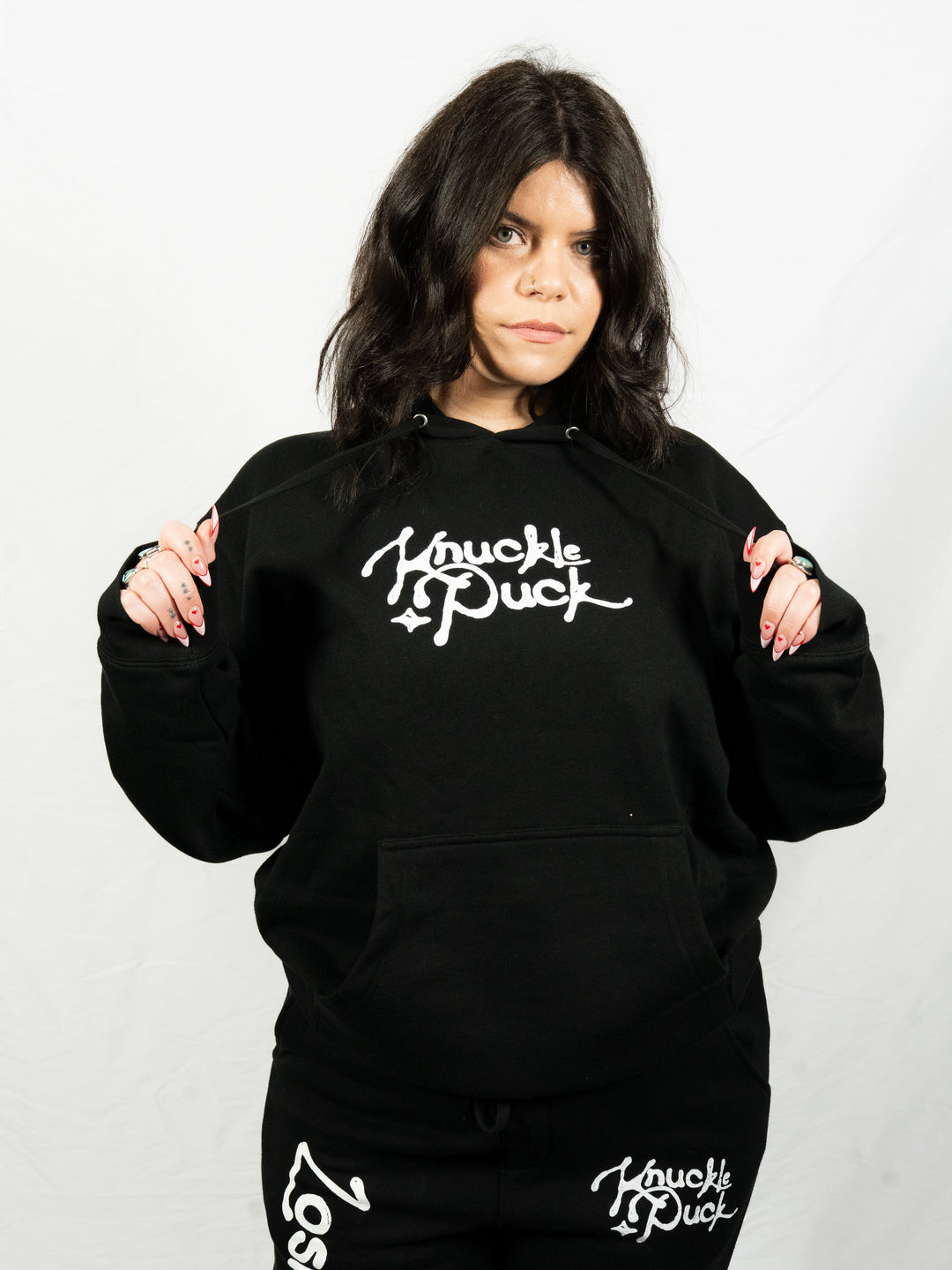 A dark-haired person in matching Knuckle Puck printed apparel—a black hoodie and pants—stands against a white background, holding the hoodie strings.