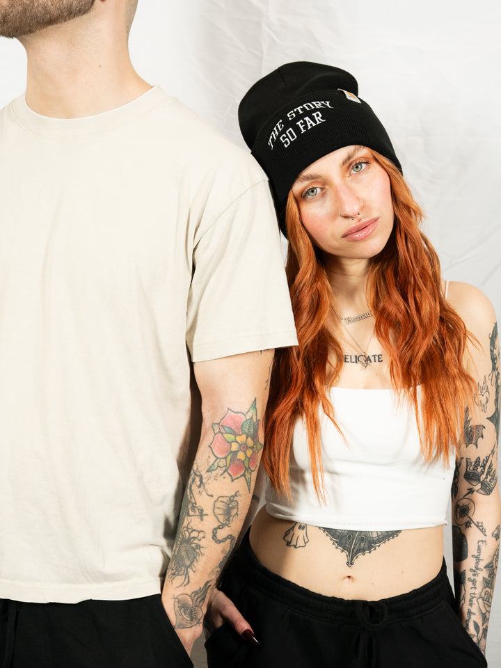 A woman with long red hair, tattoos, and a black embroidered "The Story So Far" beanie rests her head on the shoulder of a man in a beige t-shirt, his face out of frame, against a plain white background.
