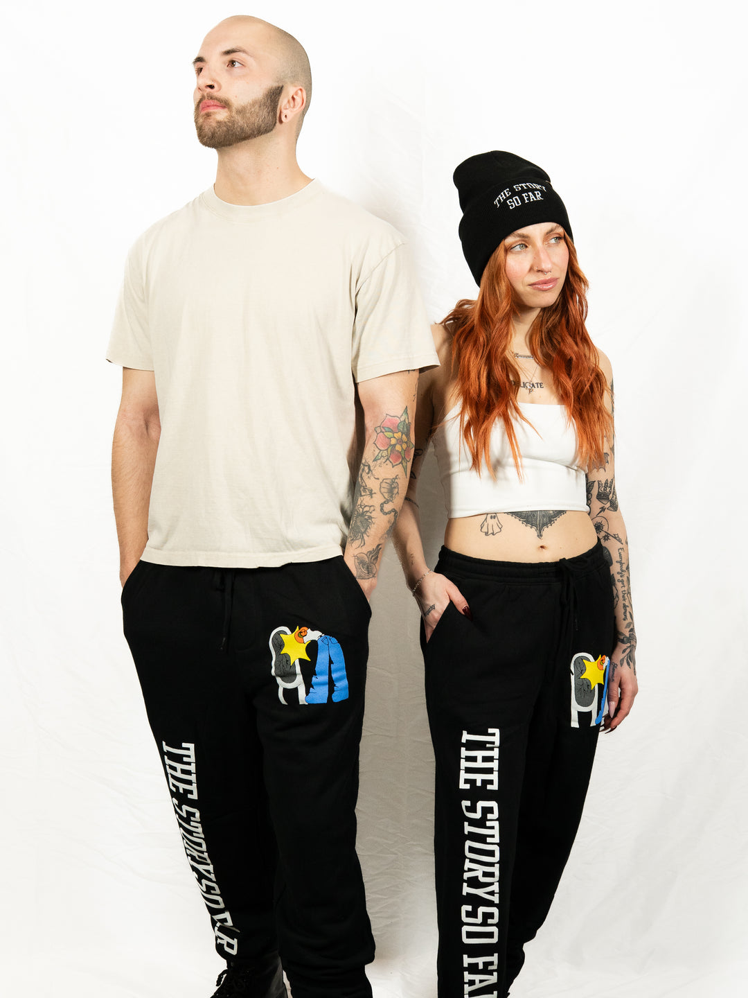 A man and woman pose against a white background wearing The Story So Far midweight fleece sweatpants with colorful designs and text. He wears a beige tee; she pairs hers with a white crop top and black beanie.