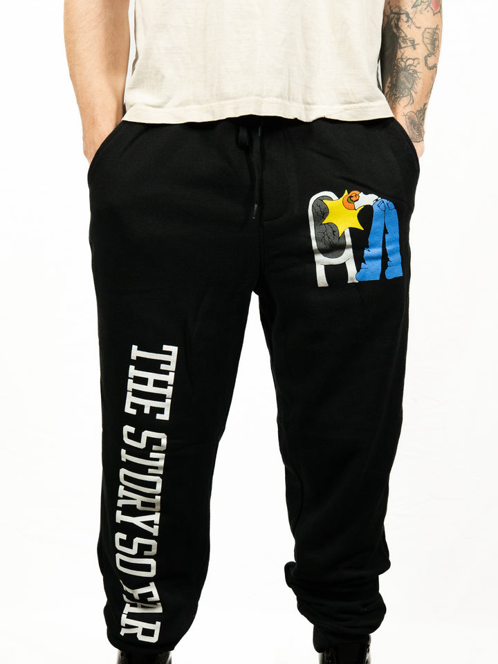 Person wearing black midweight fleece sweatpants from The Story So Far, featuring the band’s name in bold white print on one leg and colorful cartoon-style PA letters on the other; hands in pockets, white shirt partly visible.