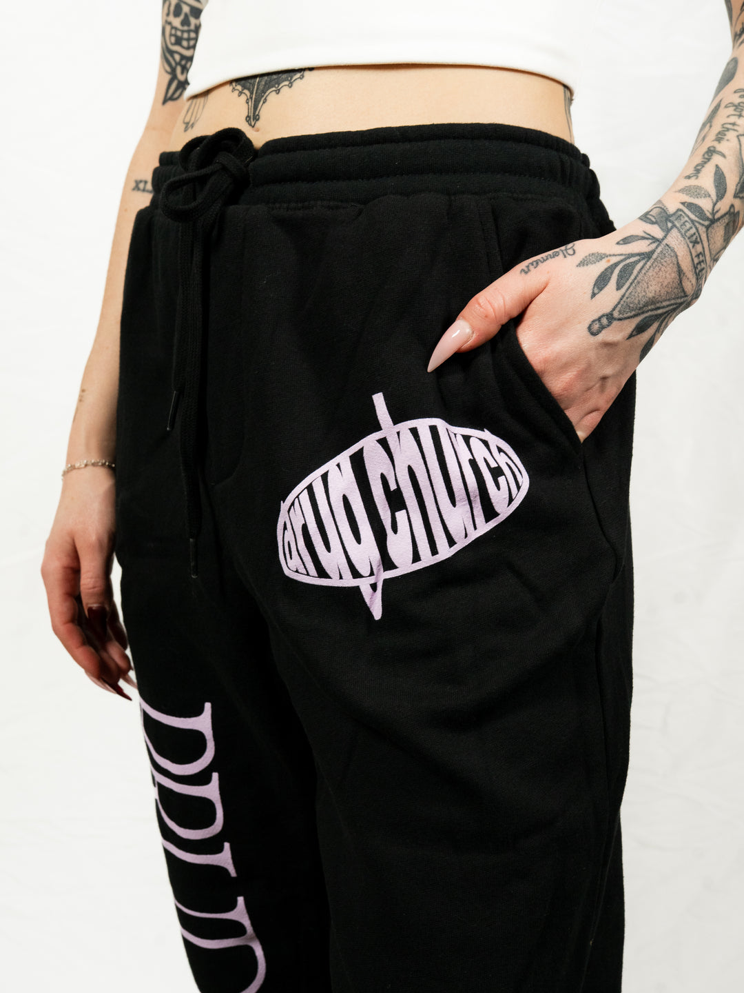 A tattooed person wears a white crop top and black Drug Church midweight fleece sweatpants with pink "drug church" lettering, one hand in their pocket.