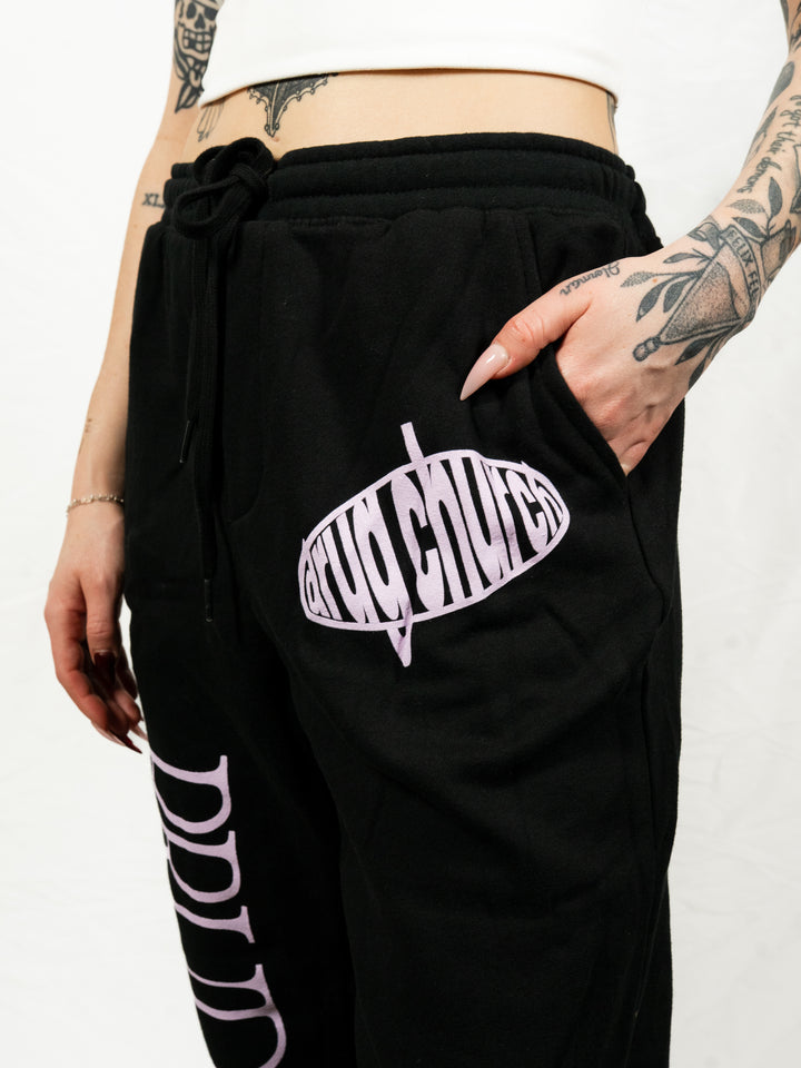 A tattooed person wears a white crop top and black Drug Church midweight fleece sweatpants with pink "drug church" lettering, one hand in their pocket.