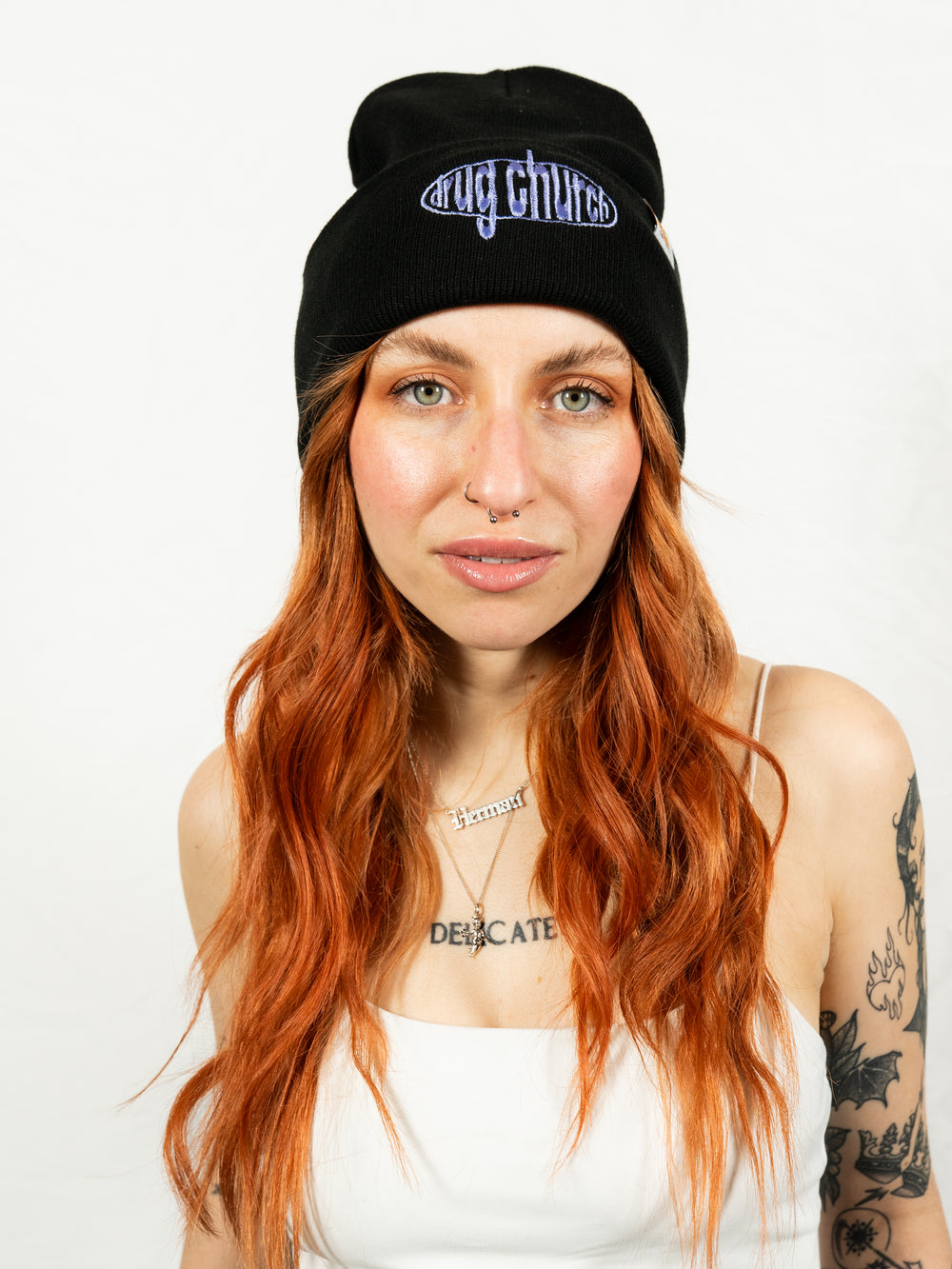 A woman with long red hair wears a black embroidered beanie featuring the Drug Church logo, a white tank top, and displays several tattoos against a plain white background. Explore unique Drug Church embroidered beanies for every style.