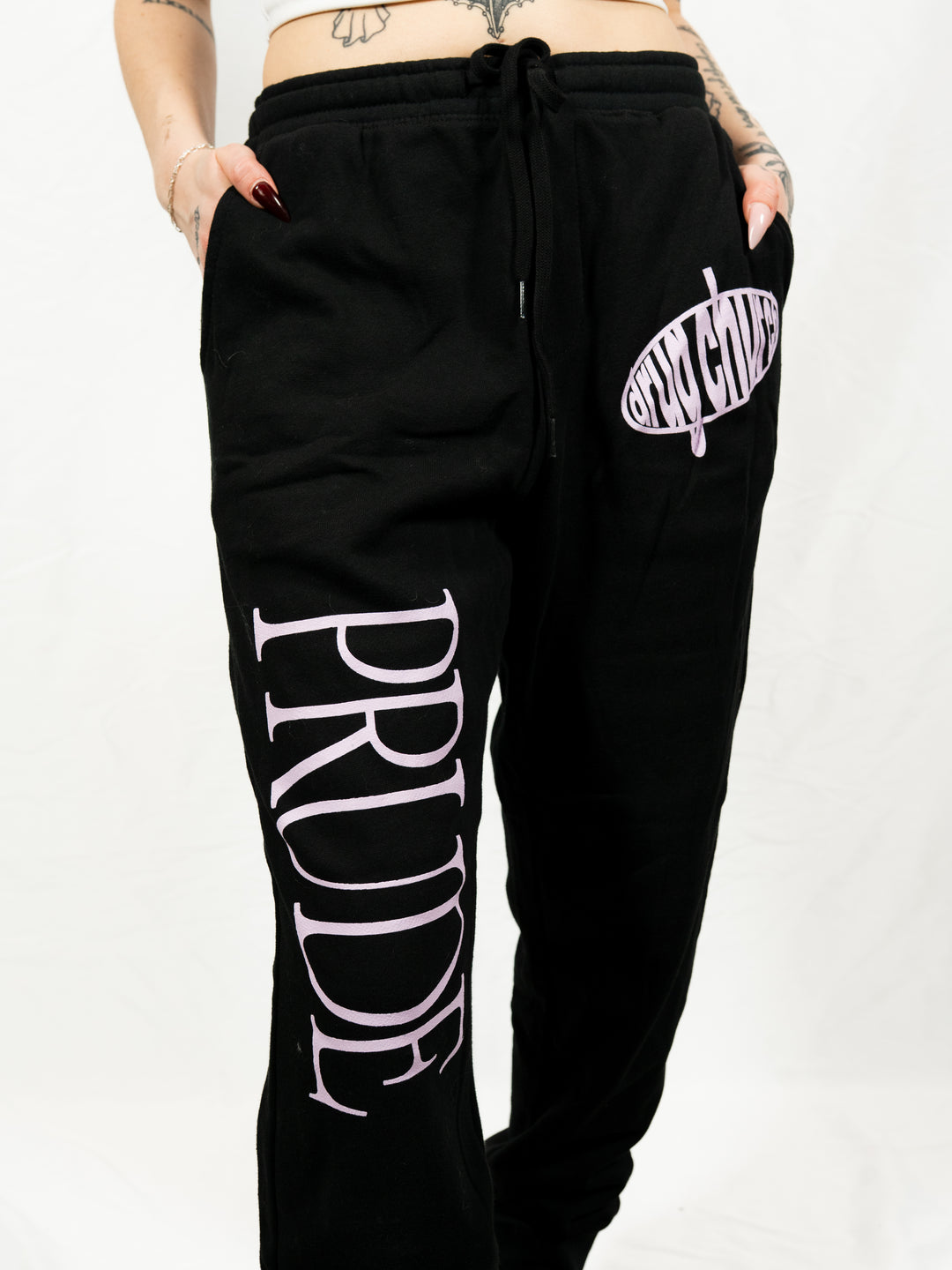 Person wearing black fleece sweatpants from Drug Church, featuring PRIDE in large pink letters down one leg and an oval-shaped pink anything logo on the other. Only the lower body is visible.