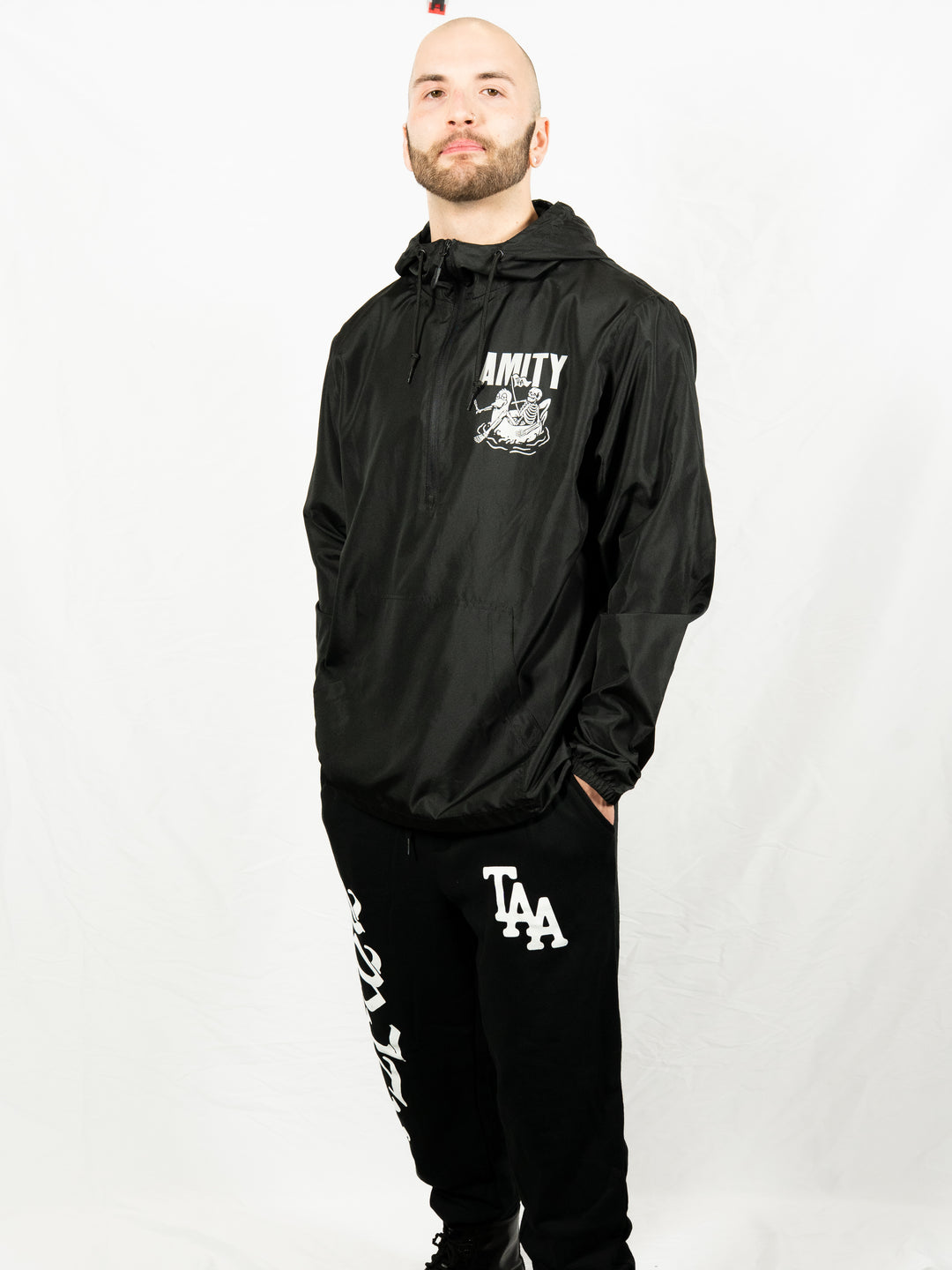 A person with a trimmed beard stands against a white background, wearing a black jacket with The Amity Affliction branding and graphic, and IND20PNT midweight fleece sweatpants by Independent Trading Co with bold white text and TAA initials on the leg.